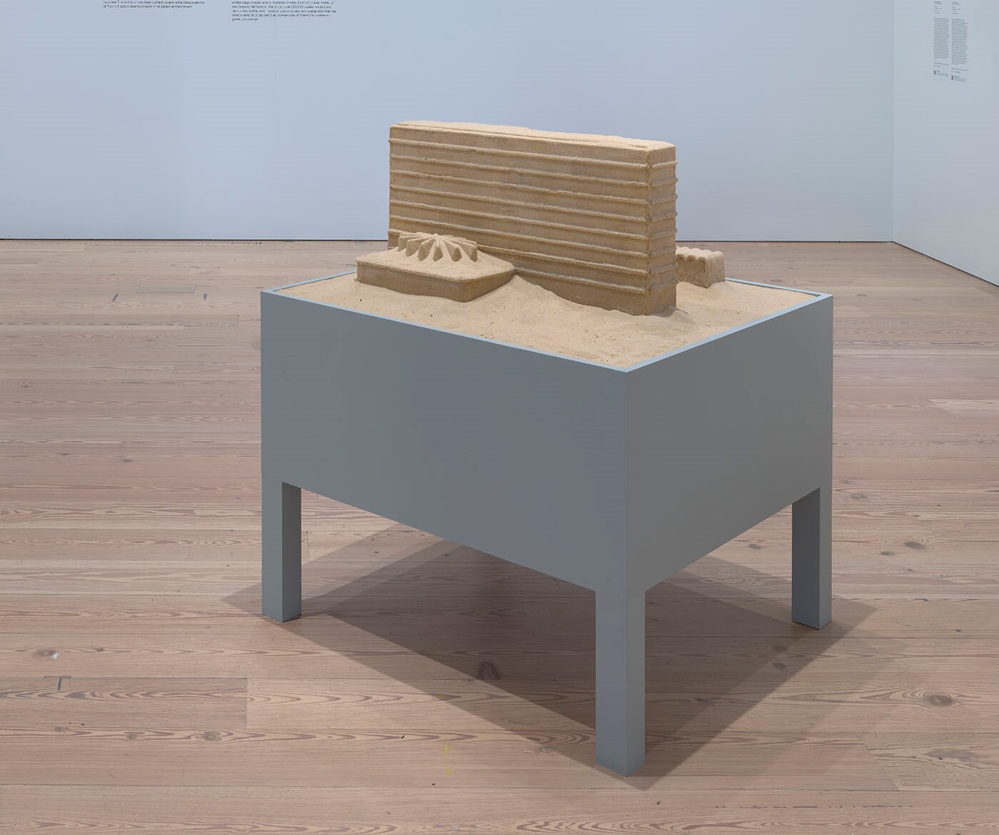 A rectangular sand sculpture with layered ridges sits on a gray pedestal in a gallery.