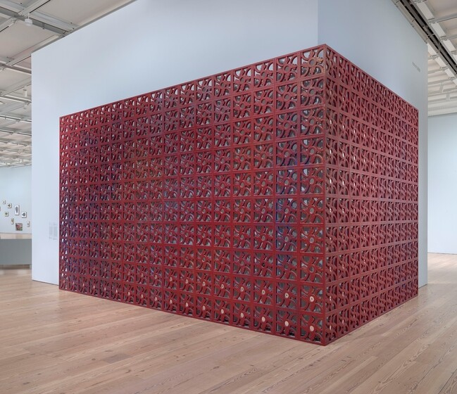 A large, red, geometric lattice sculpture stands prominently in a modern art gallery with white walls and wooden floors. The intricate pattern of the sculpture contrasts with the minimalist surroundings, and a series of framed artworks can be seen in the background to the left.