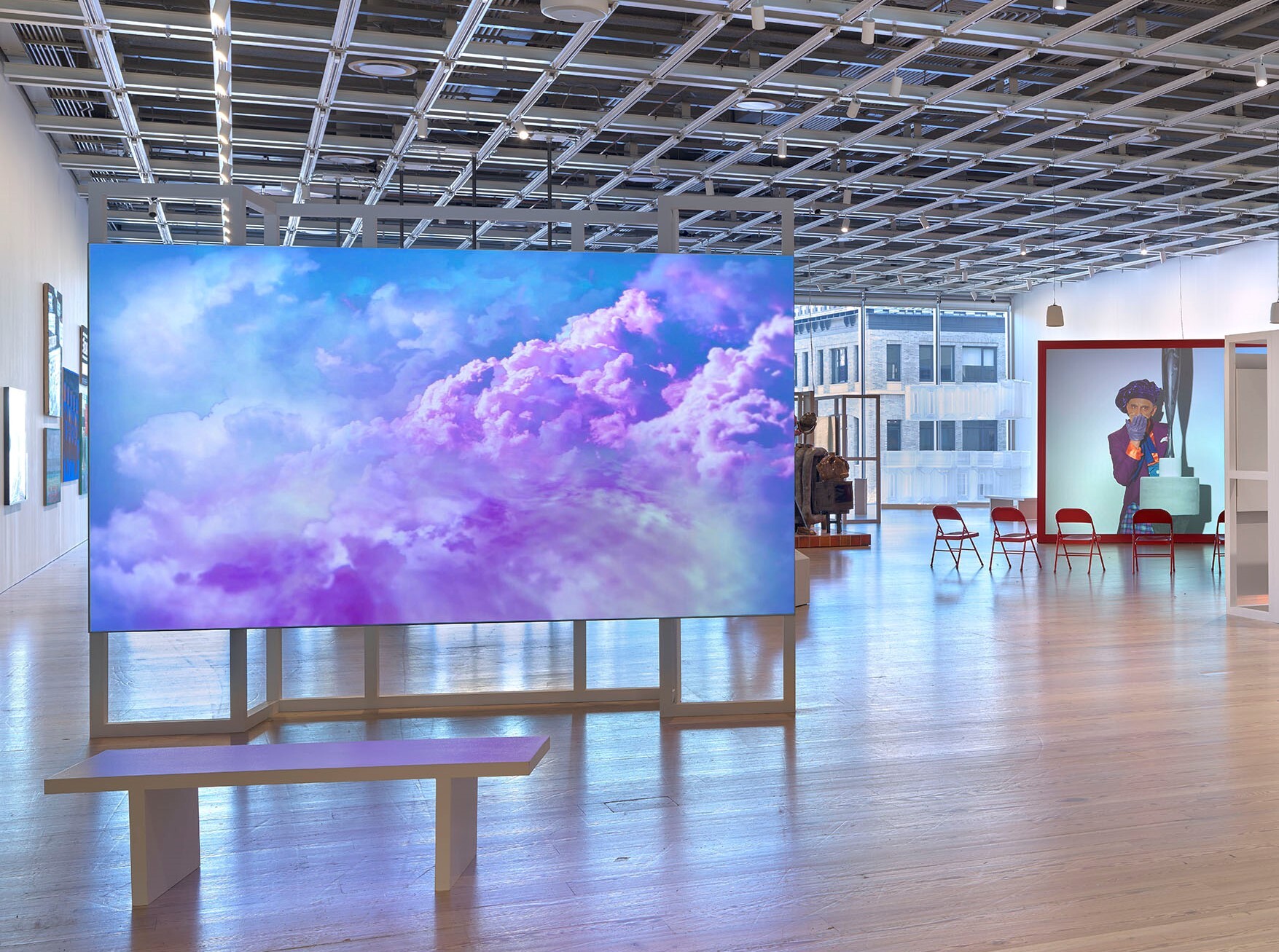 A spacious art gallery with a large, vibrant digital display of purple and blue clouds on a central screen. The gallery features a high ceiling with industrial lighting, wooden floors, and a bench for viewing the artwork. In the background, there are more artworks, red chairs, and a sculpture, with large windows allowing natural light to fill the space.