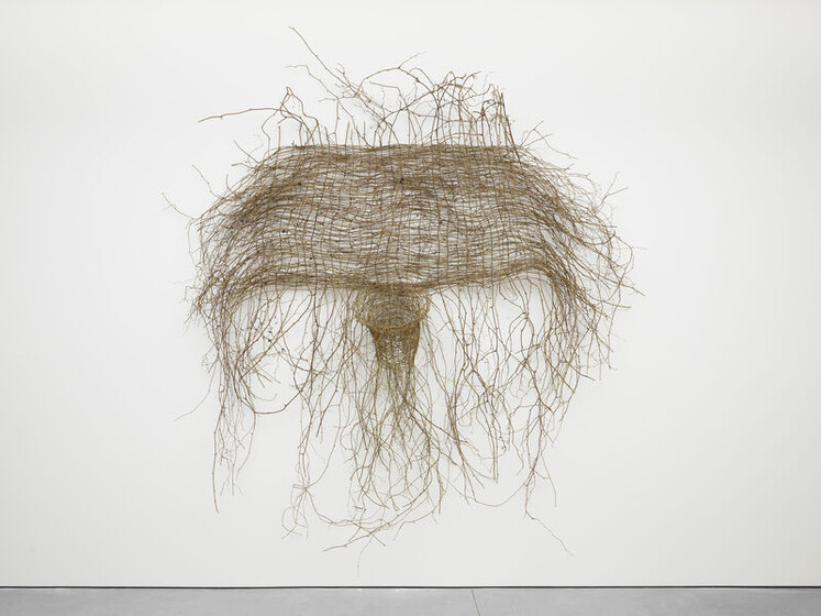 A sculptural artwork consisting of a dense network of interwoven branches or twigs suspended in mid-air against a white gallery wall, with numerous tendrils hanging down towards the floor.