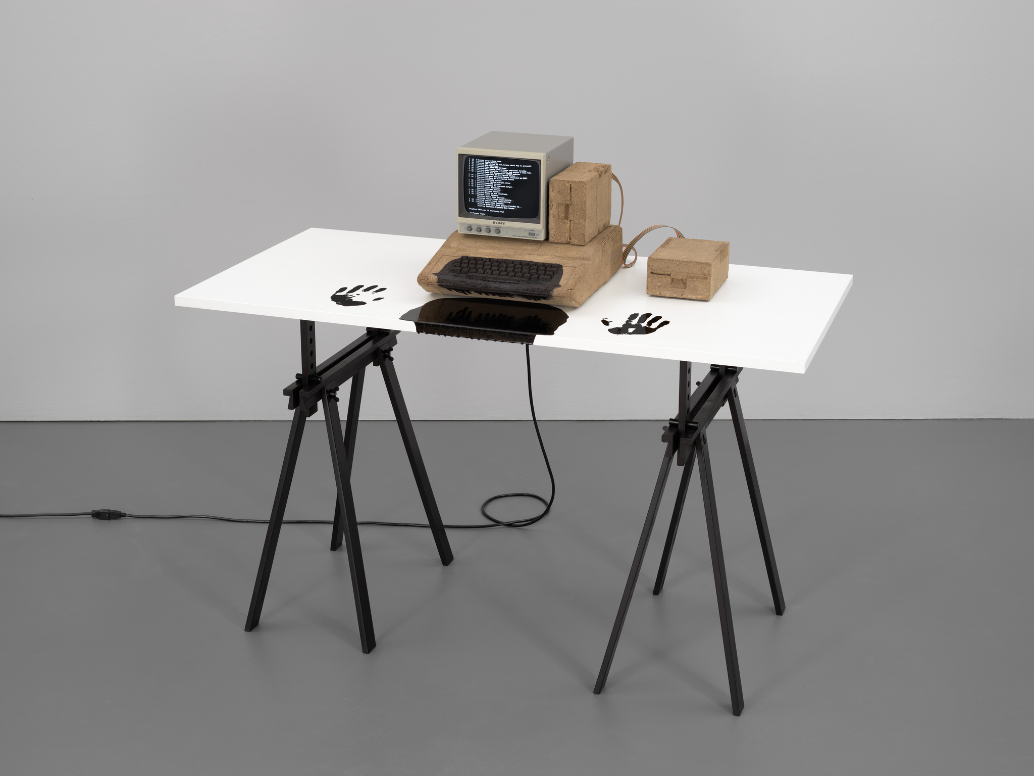 A computer made mostly of dirt with asphalt pooling on the keyboard and desk and two black handprints on either side.