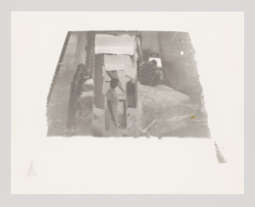 A person standing in a narrow hallway between beds and curtains in a dim room.