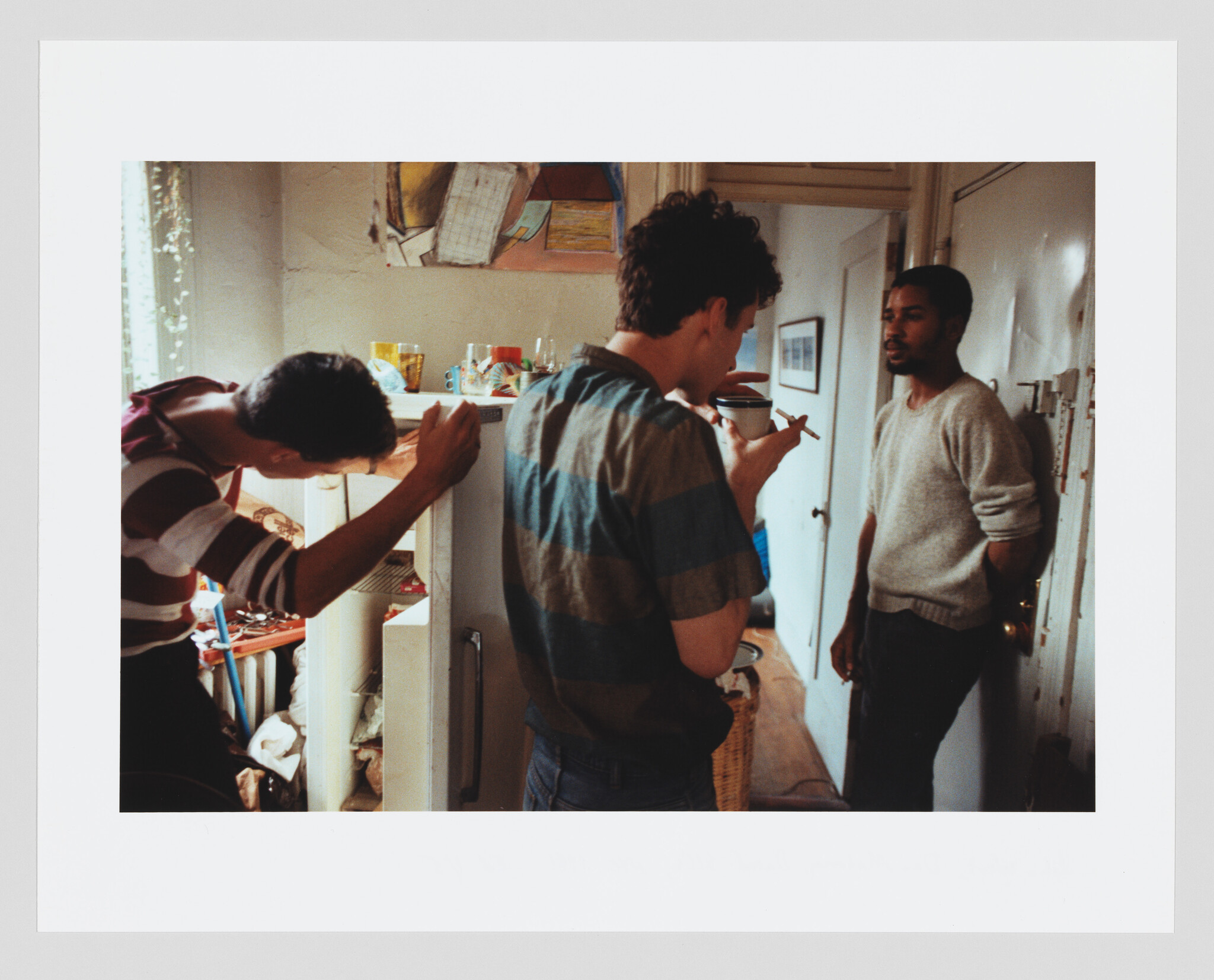 Three people in a small kitchen, one looking in the fridge, another drinking coffee, and one standing by the door.