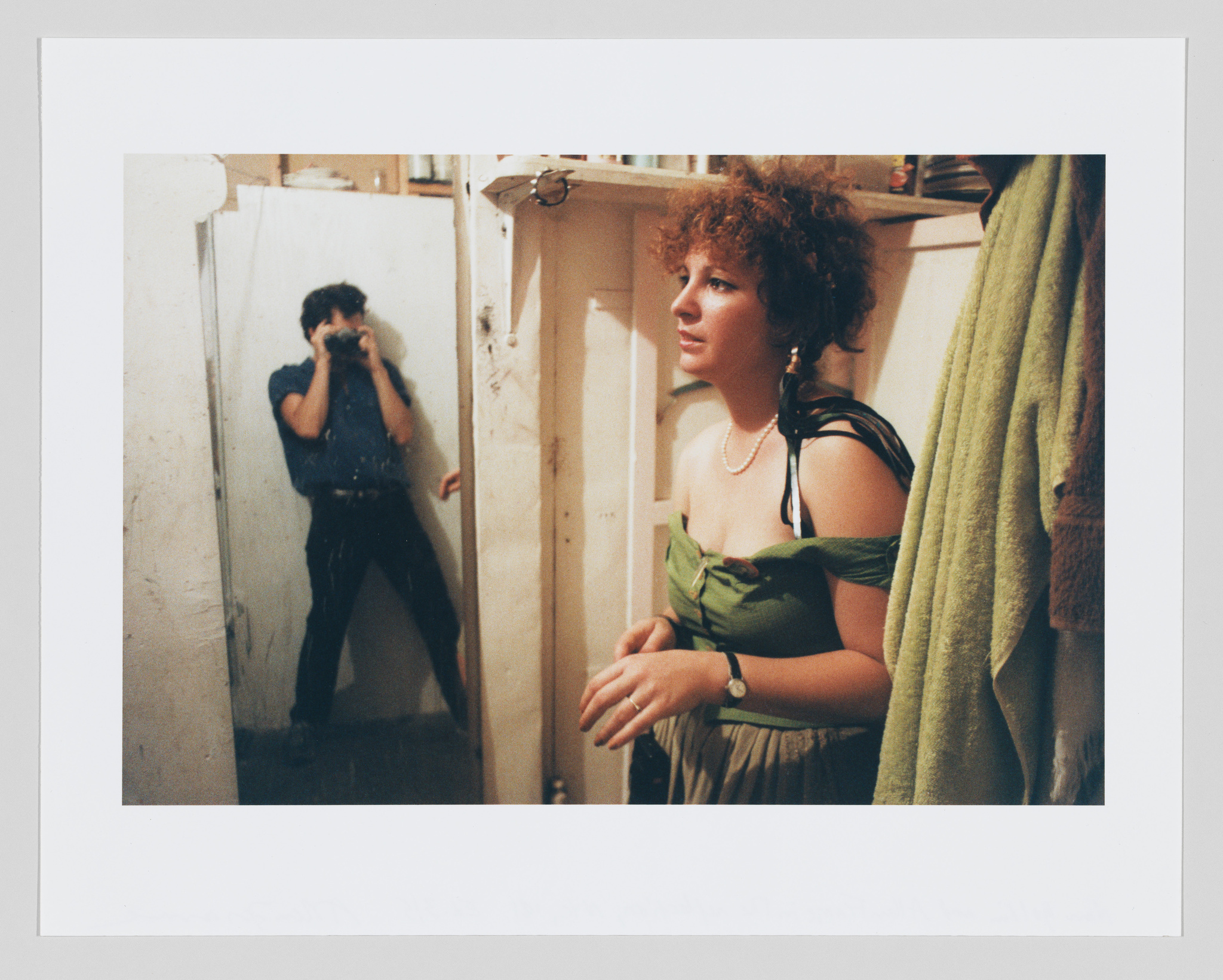 A photograph capturing a candid moment where a man is taking a picture of himself in a mirror, while a woman in a green dress stands to the side, looking away with a thoughtful expression. The setting appears to be a small, cluttered room with a towel hanging nearby.