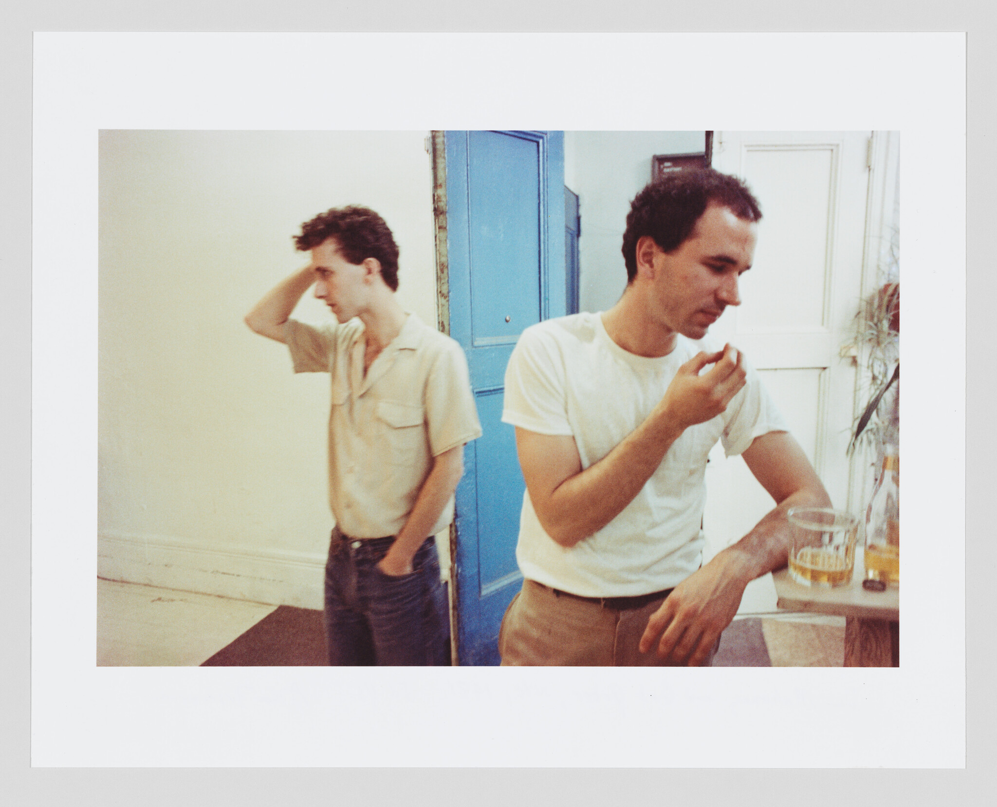 Two men in a room with vintage decor; one is standing in profile with his hand on his head, appearing contemplative, while the other is seated at a table, eating with a glass in front of him. The room has a blue door and trim, with a white wall and a plant in the background.