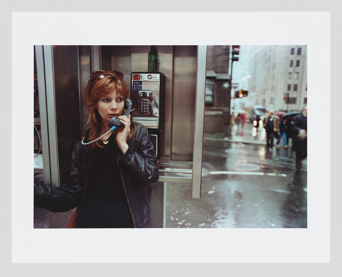 A woman with red hair is standing in a phone booth, speaking into the receiver. She's wearing a black leather jacket and has sunglasses perched on her head. The scene outside the booth shows a rainy city street with pedestrians carrying umbrellas.