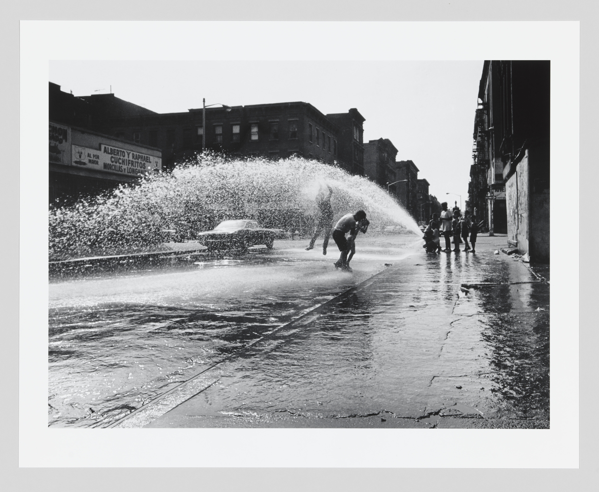 Children play in a street with an open fire hydrant spraying water, in a black and white urban scene.