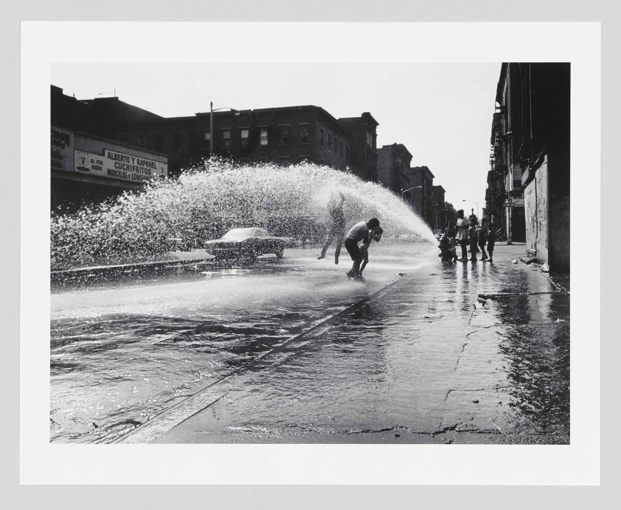 Children play and run through a large spray from an opened fire hydrant on a city street.