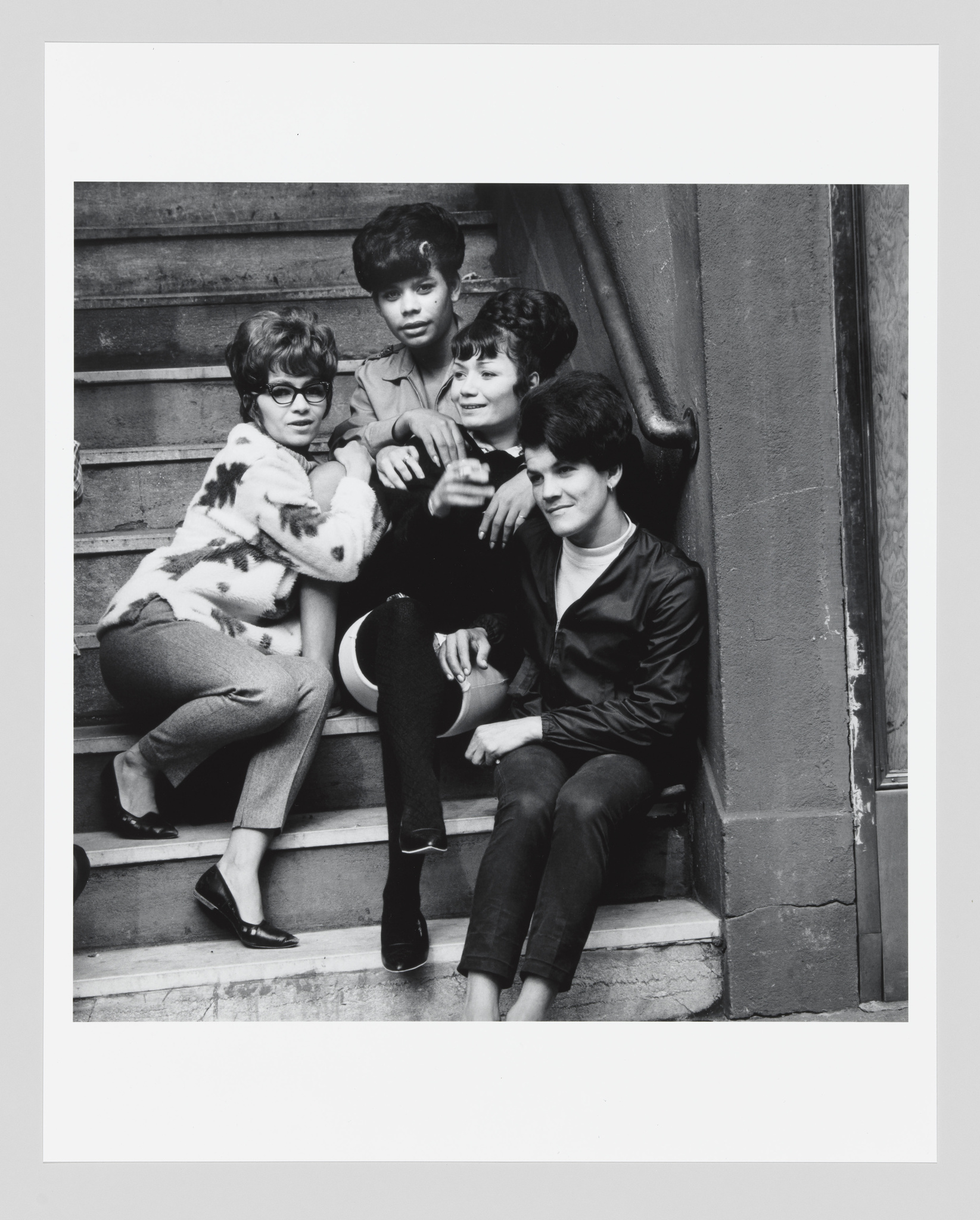 A black and white photo of four young women sitting closely together on outdoor steps, smiling and posing for the camera. They are dressed in styles that suggest the photo was taken in the mid-20th century.