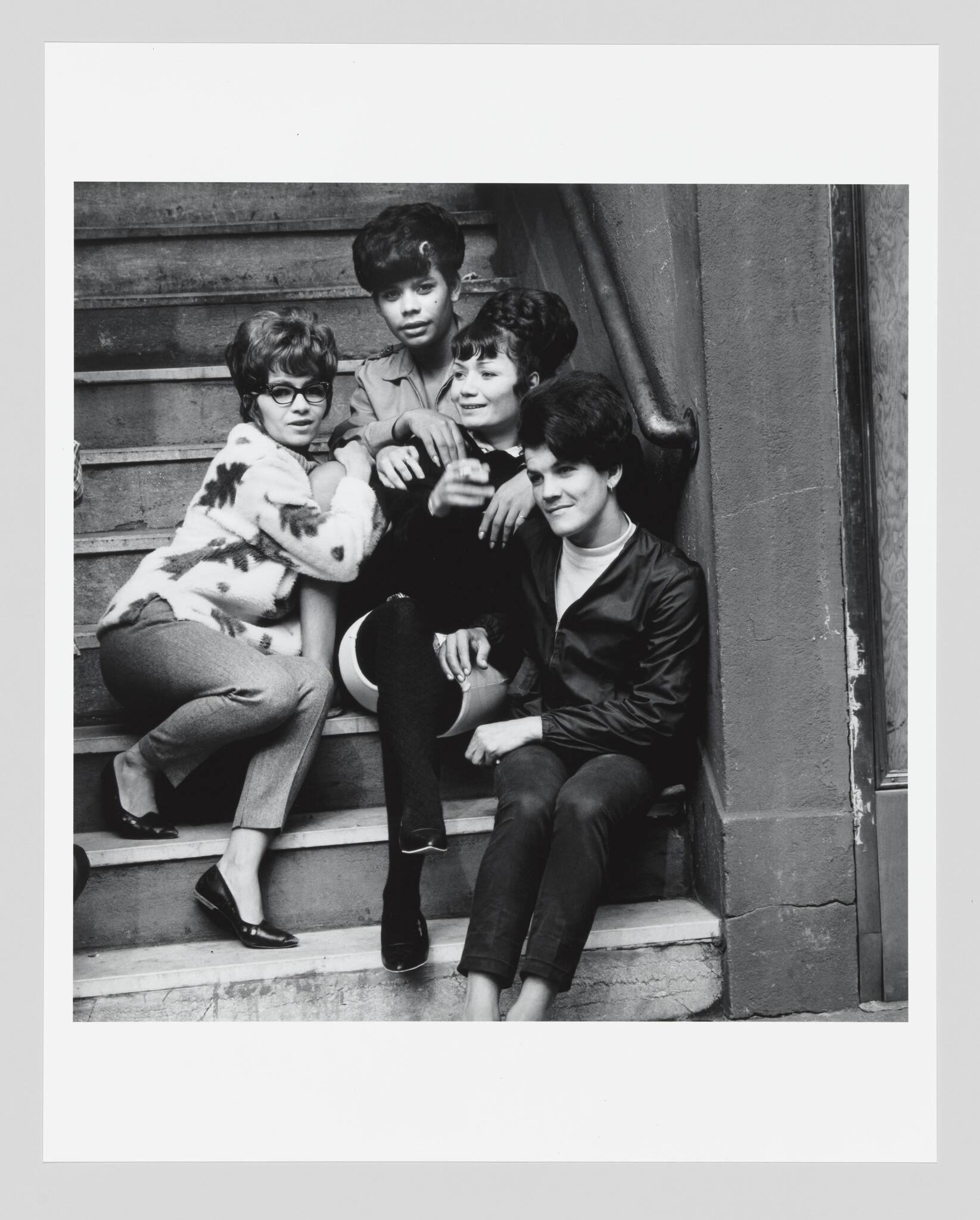 Four young women sit closely together on outdoor steps, smiling and leaning against a railing.