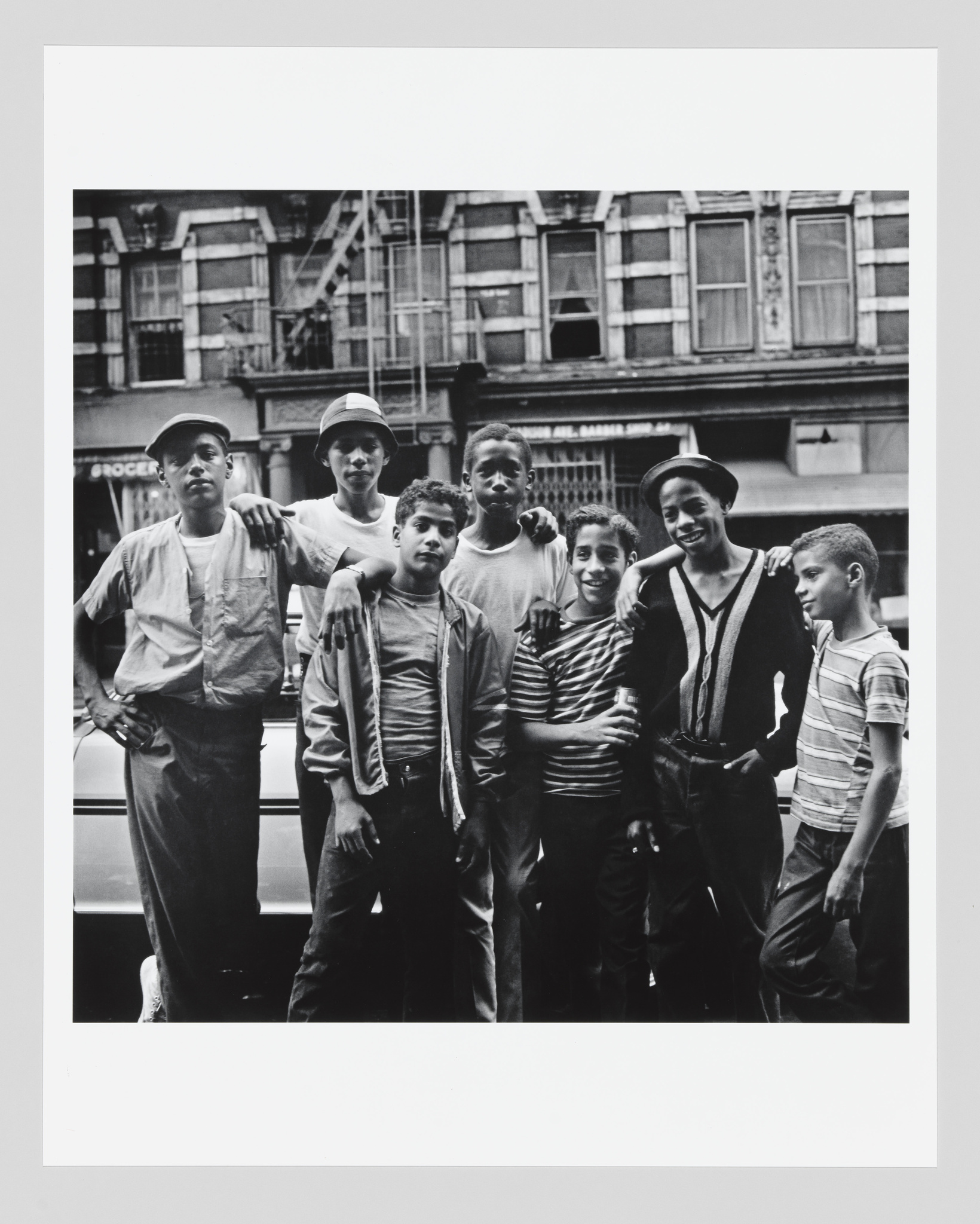 A black and white photograph of seven young boys posing together on a city street, with old buildings and a parked car in the background. They are dressed in casual clothing from an earlier era, with some wearing hats, and they exhibit a range of expressions and poses, suggesting camaraderie and youthful confidence.