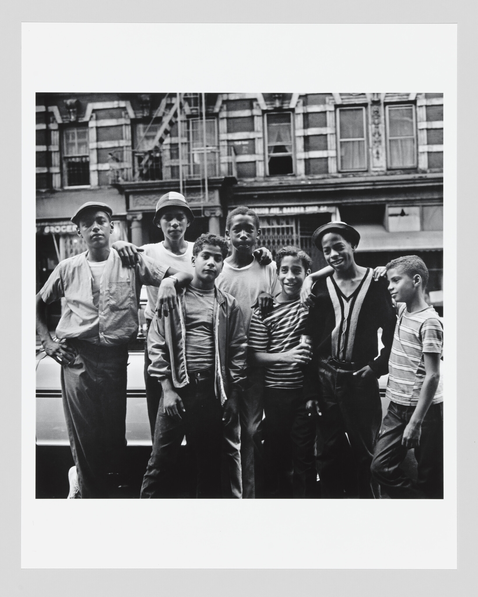 Seven boys stand together on a city sidewalk with arms around each other's shoulders.
