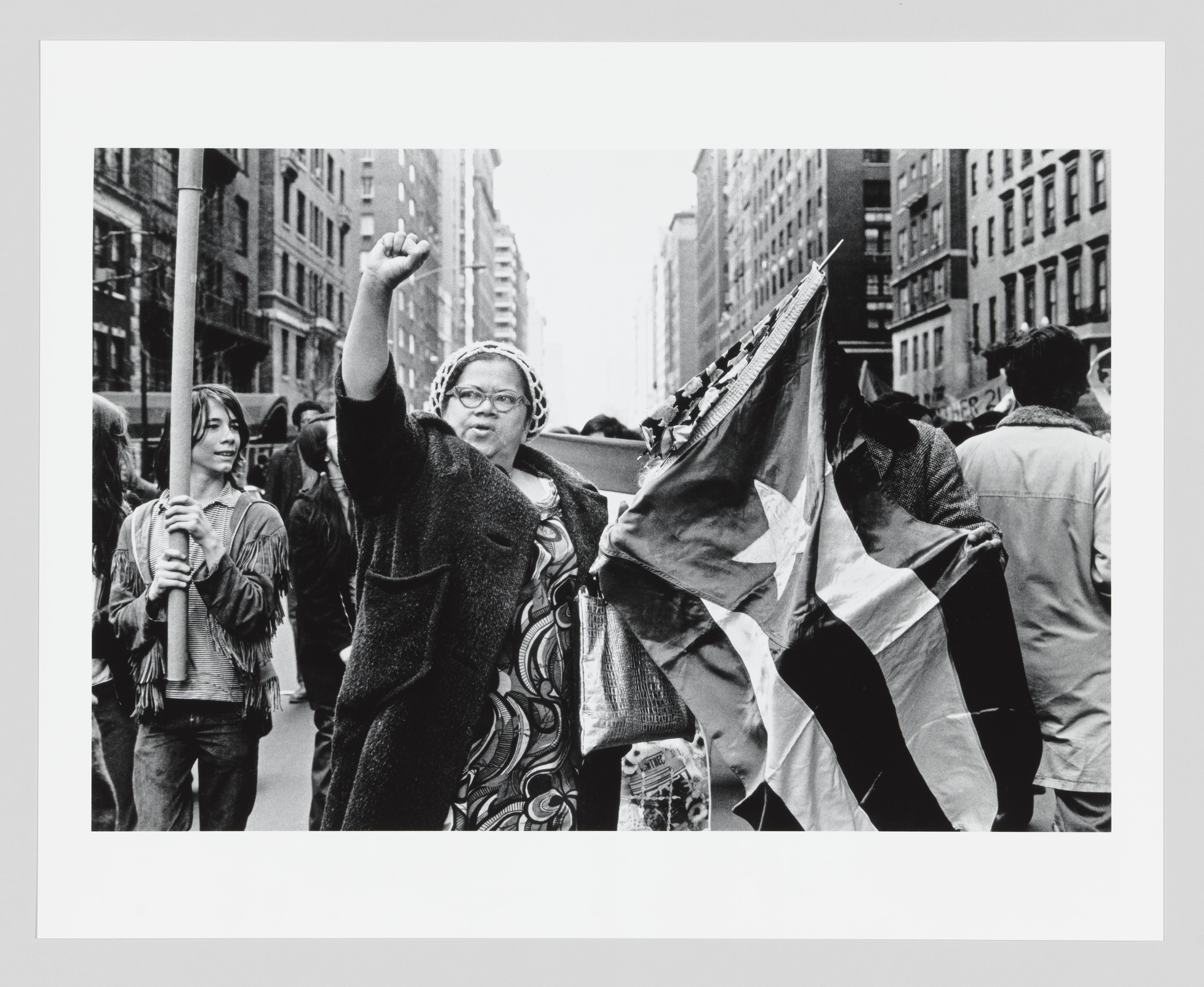 A black and white photograph capturing a moment during a protest or public gathering. A woman in the center raises her fist high, wearing glasses and a patterned dress, conveying a sense of determination or solidarity. To her left, another individual holds a pole with a tattered flag, while various other participants can be seen in the background, suggesting a lively and possibly charged atmosphere.