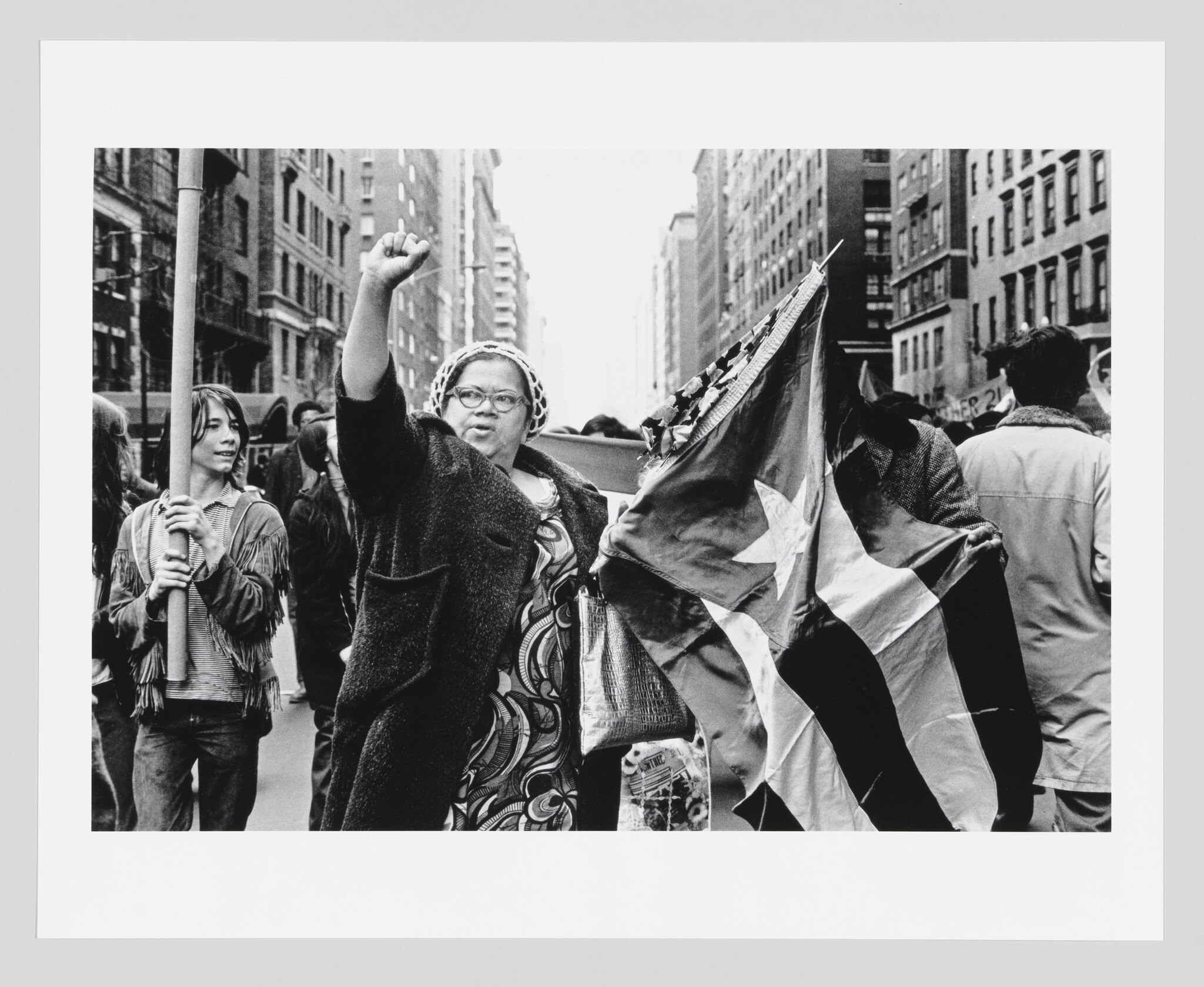 Woman raises her fist and holds a large star-striped flag while marching down a city street.