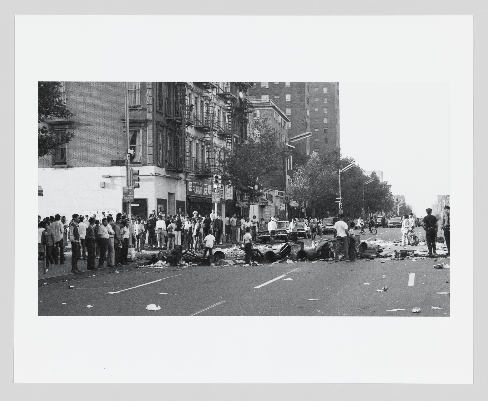 Crowd stands around a debris-filled street with overturned trash cans blocking the roadway.