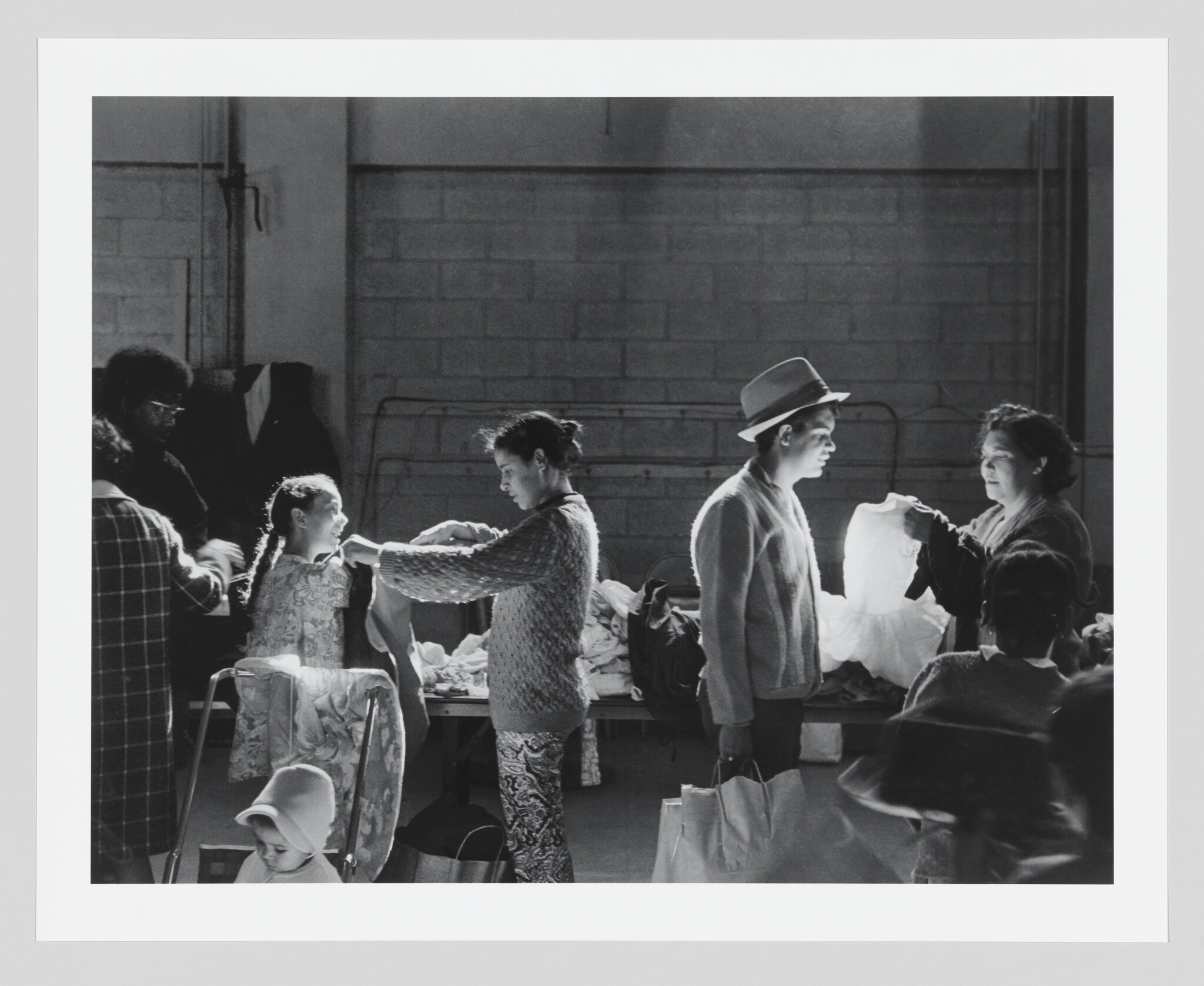 A black and white photograph capturing a candid moment backstage, where individuals are engaged in various activities, likely related to costume fitting or preparation for a performance. A woman in the center adjusts a garment on a young girl, while another woman to the right, wearing a hat, converses with someone holding up a piece of fabric. The setting appears to be an informal dressing area with a brick wall in the background and natural light streaming in from the side.