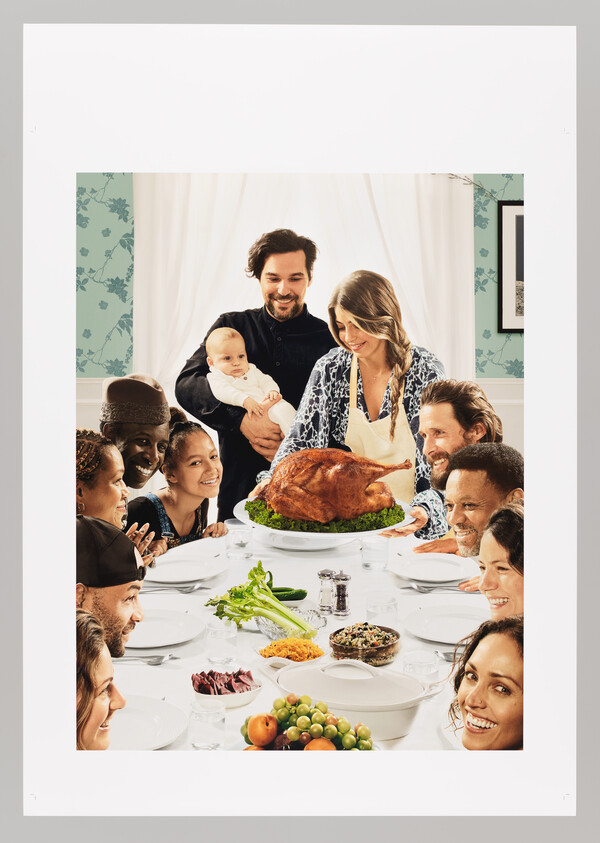 A family gathers around a table as a woman carries a roasted turkey toward smiling relatives.