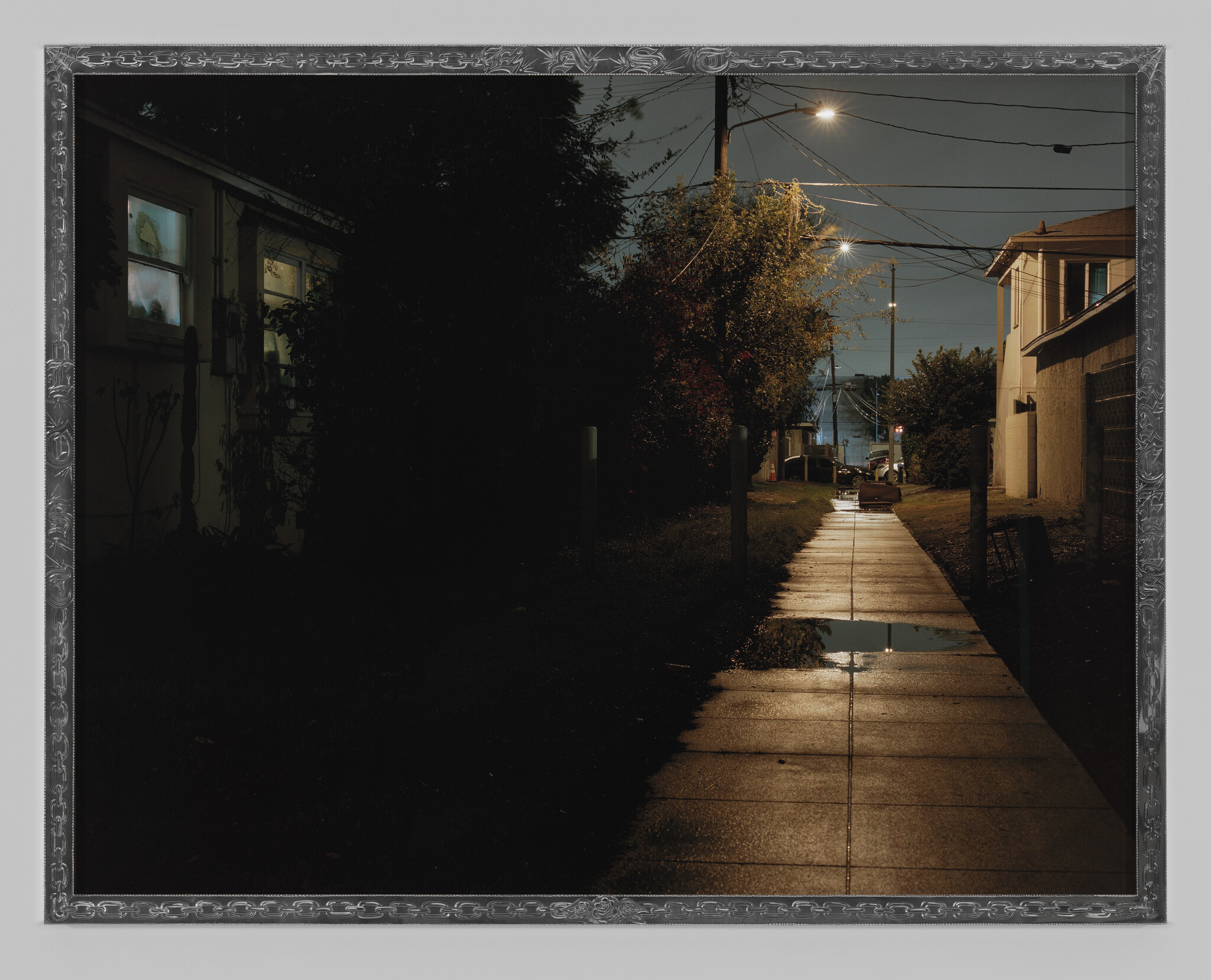A wet residential sidewalk at night reflects streetlights leading toward distant houses.