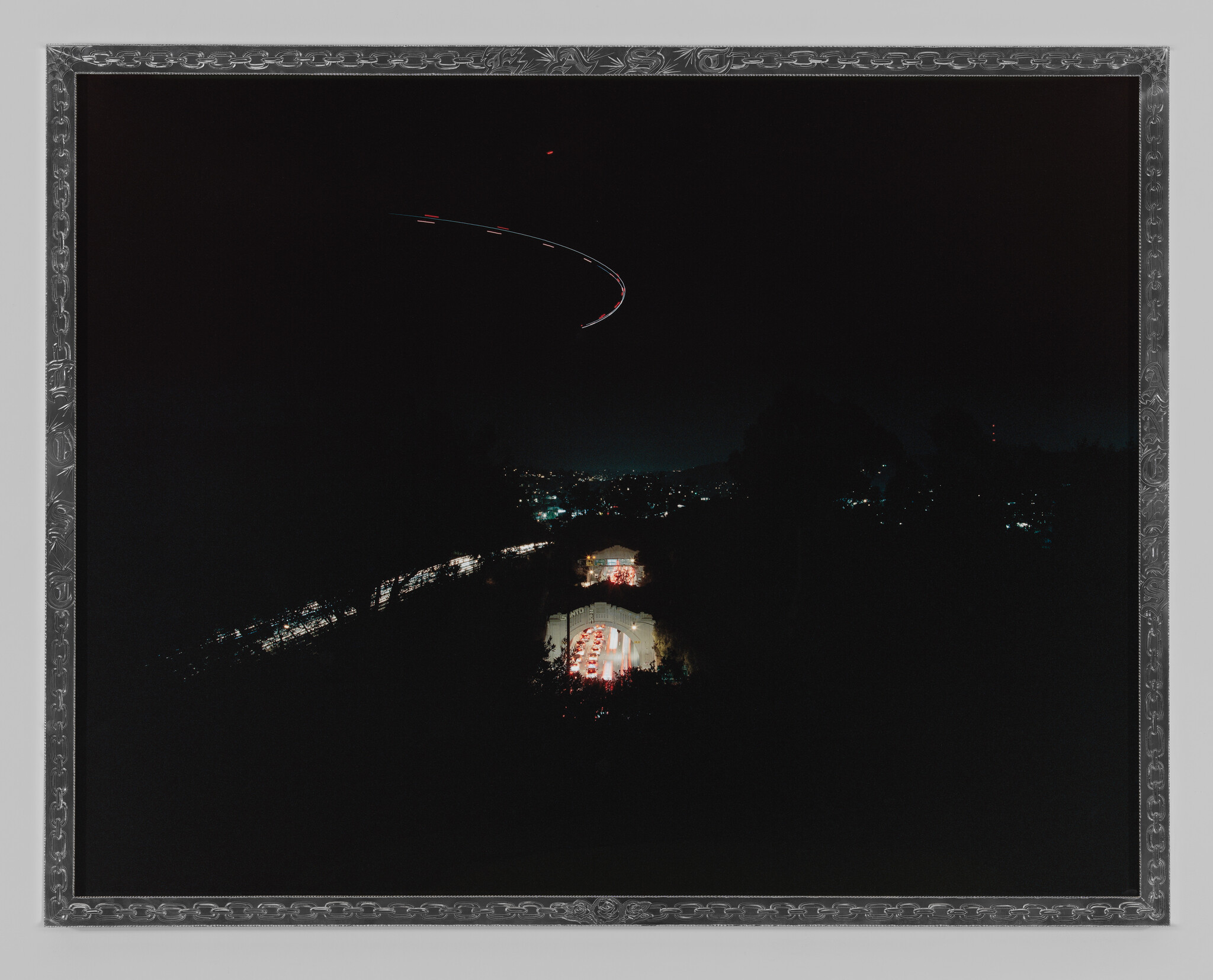 Distant illuminated arch and buildings below a curved aircraft light trail across the night sky.