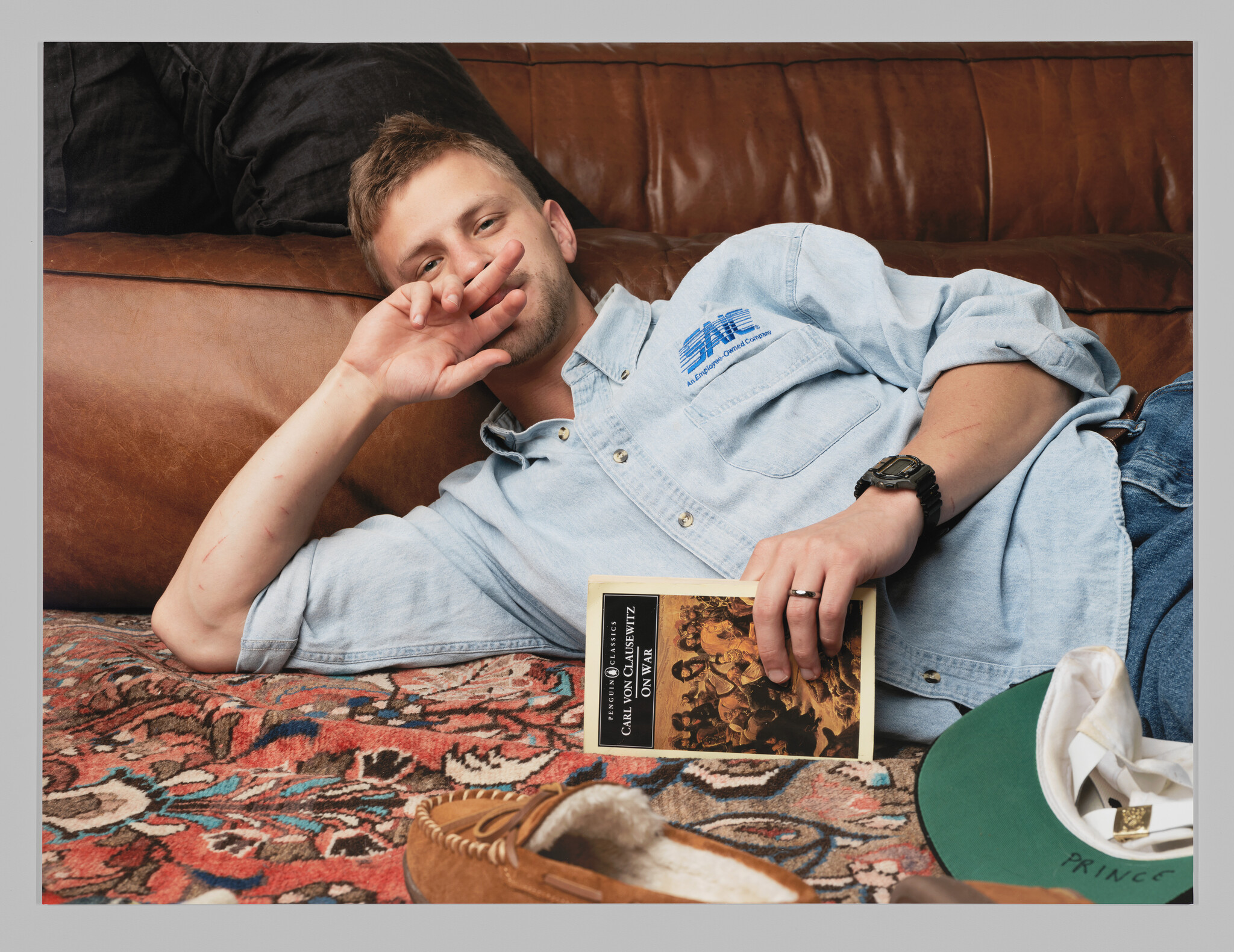 Young man lying on a couch holds a paperback book while making a peace sign toward the camera.
