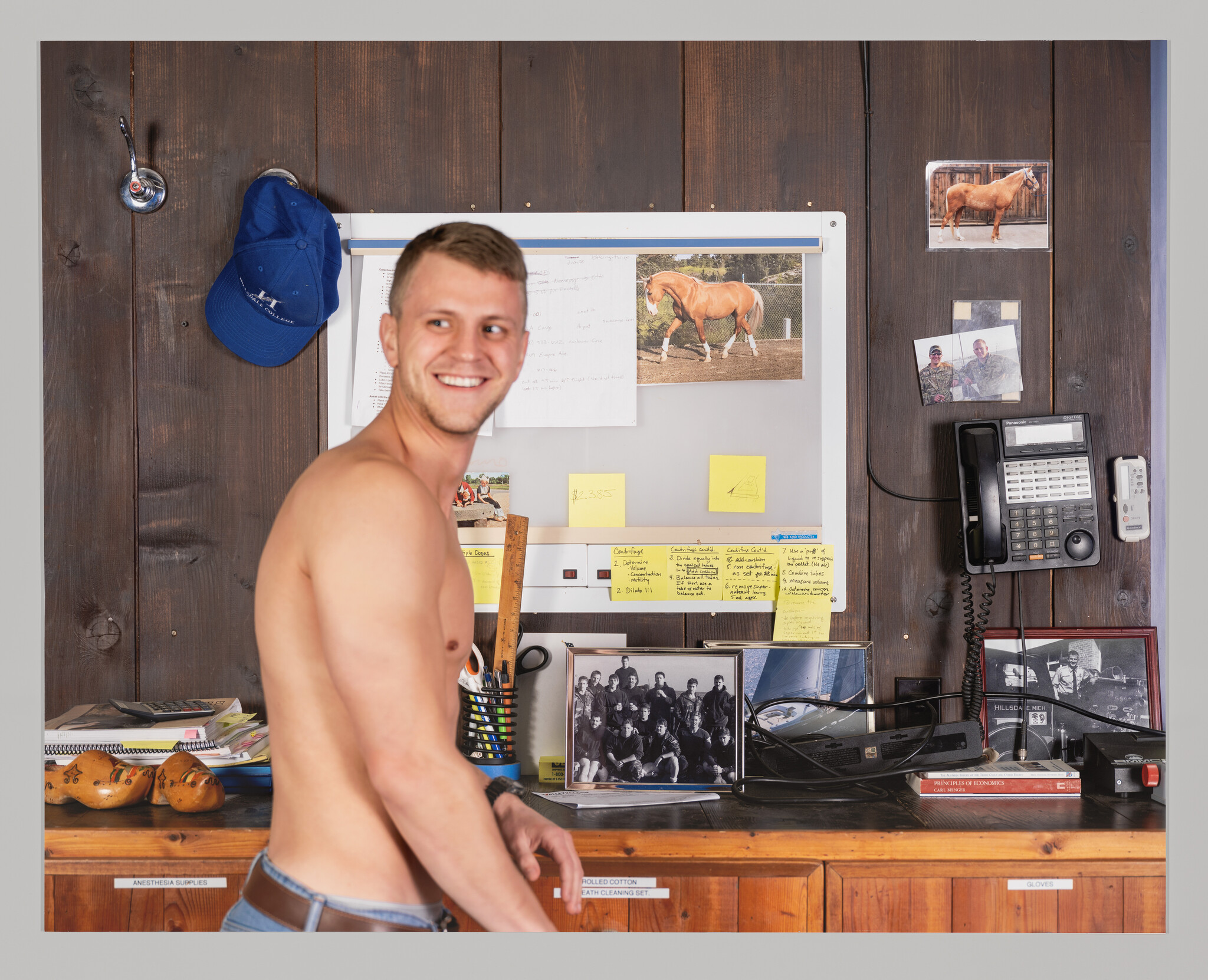A shirtless young man smiles and turns toward the camera in a cluttered office workspace.