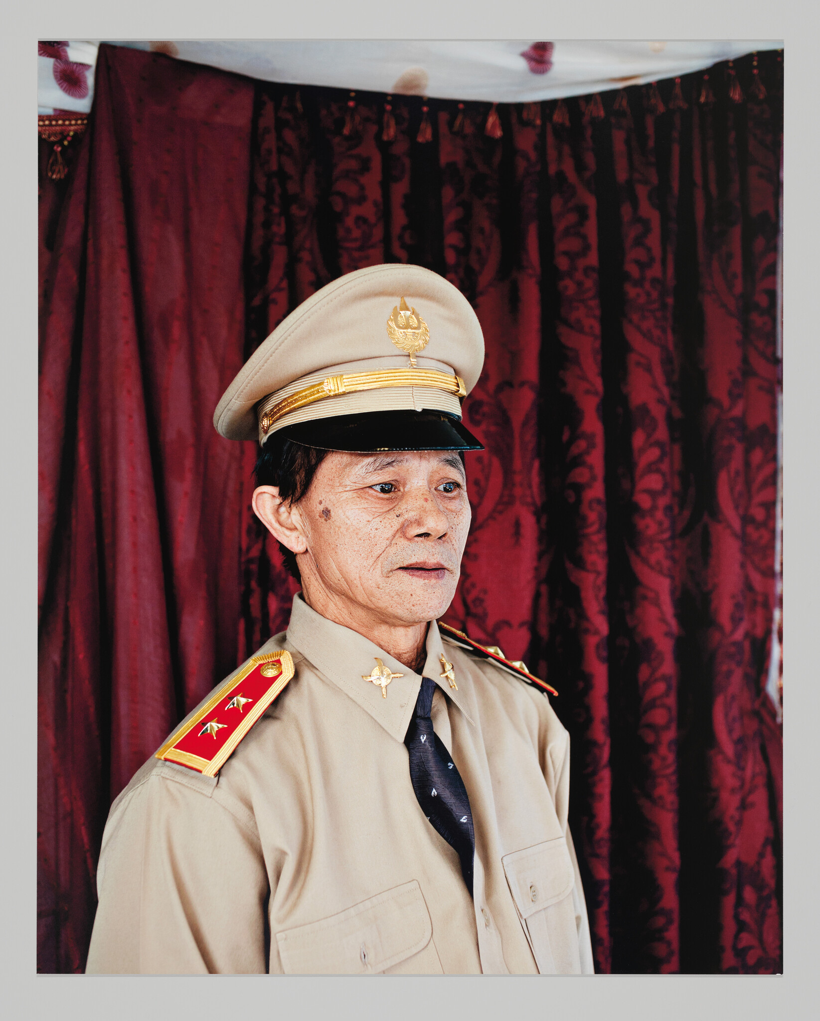 An older man wearing a beige military uniform and peaked cap stands solemnly against a red curtain.