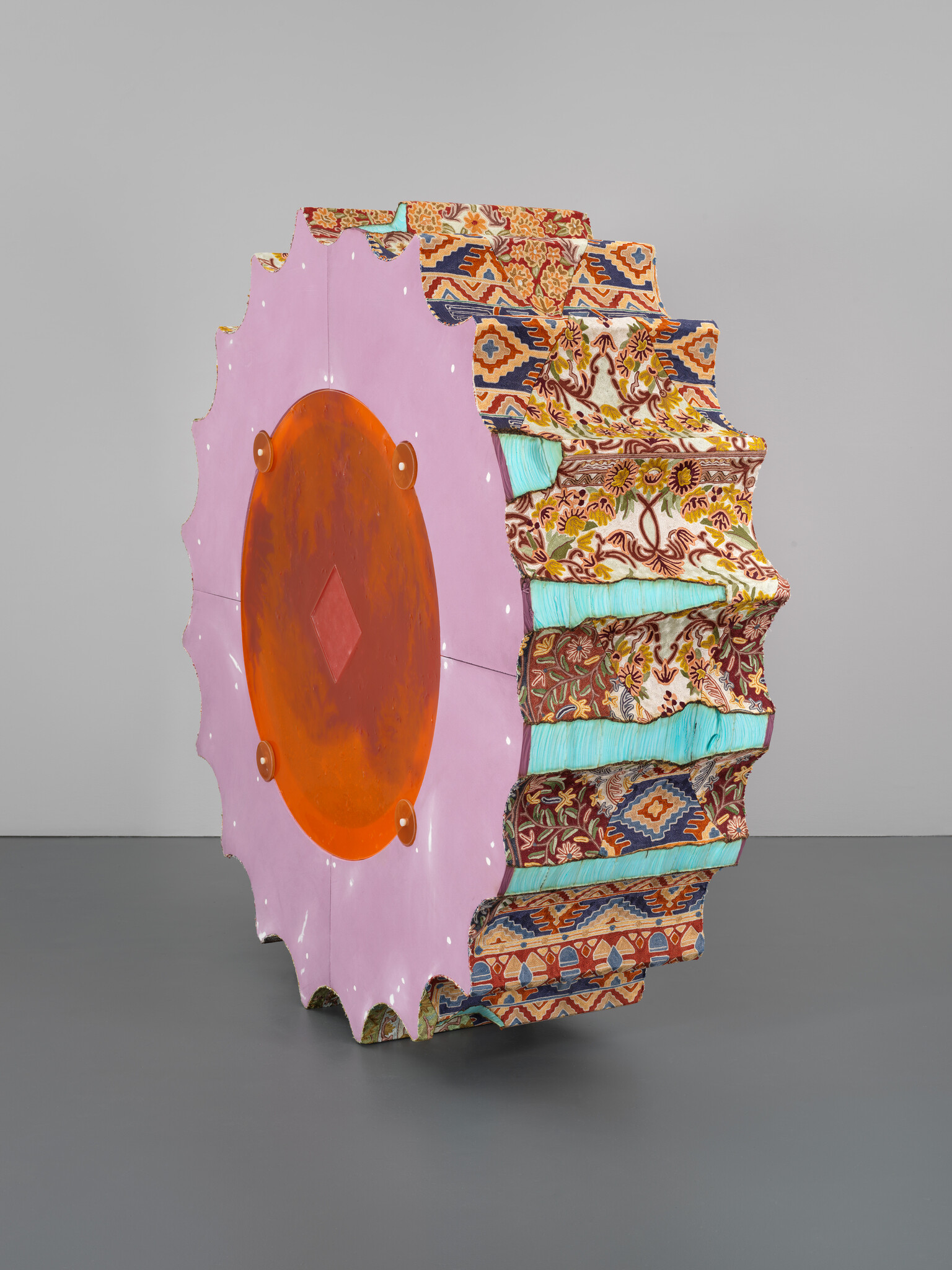 Colorful, patterned wheel sculpture with a pink and orange circular center, displayed against a gray background.