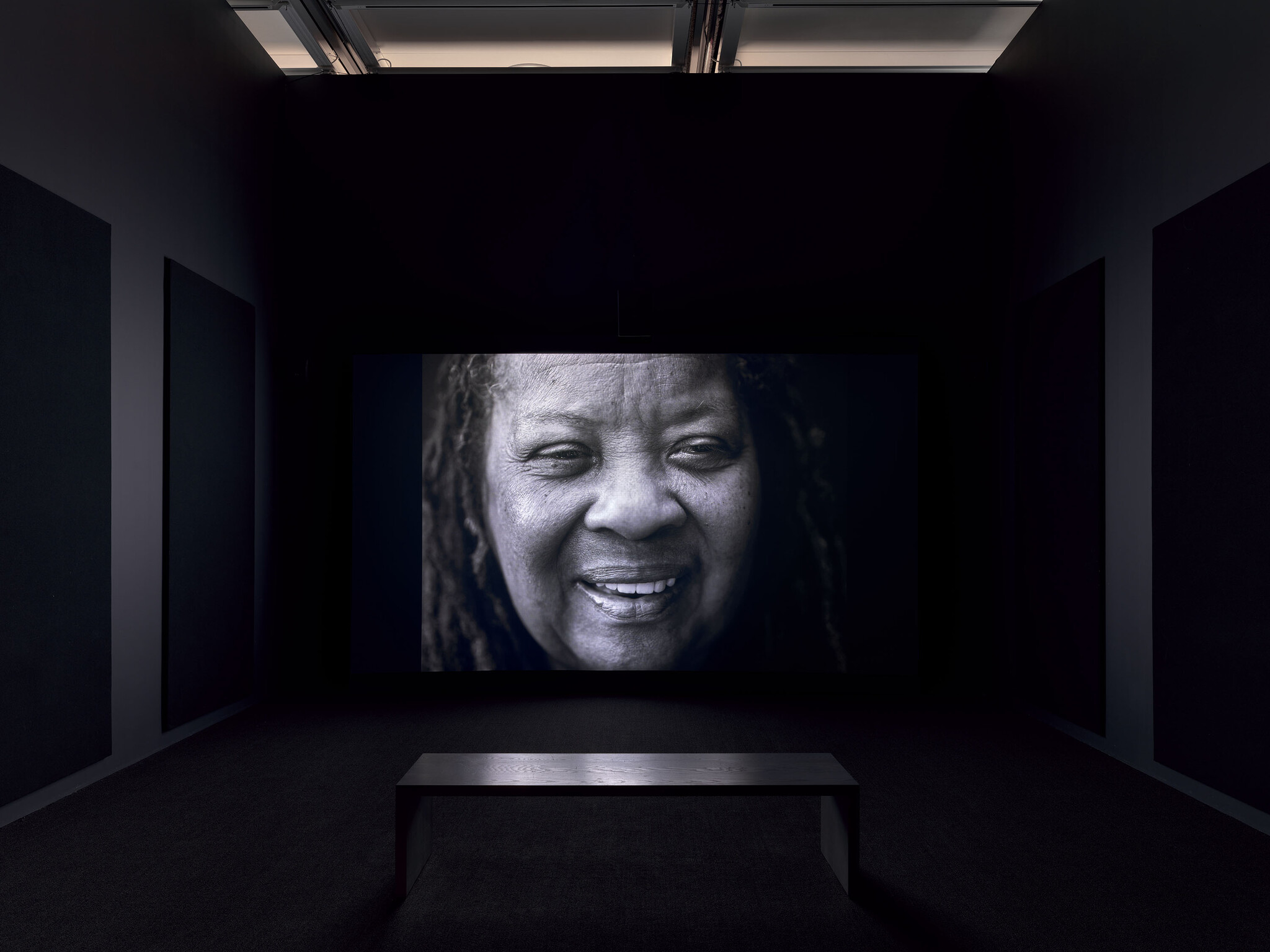 Large black-and-white close-up of a smiling older woman displayed on a dark gallery screen.