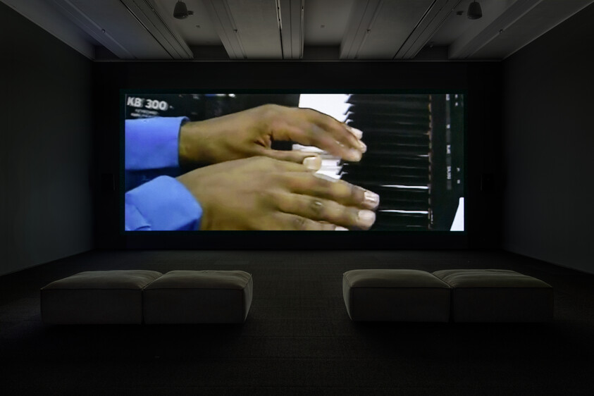 Large screen shows hands playing a keyboard, with two benches in a dark room.