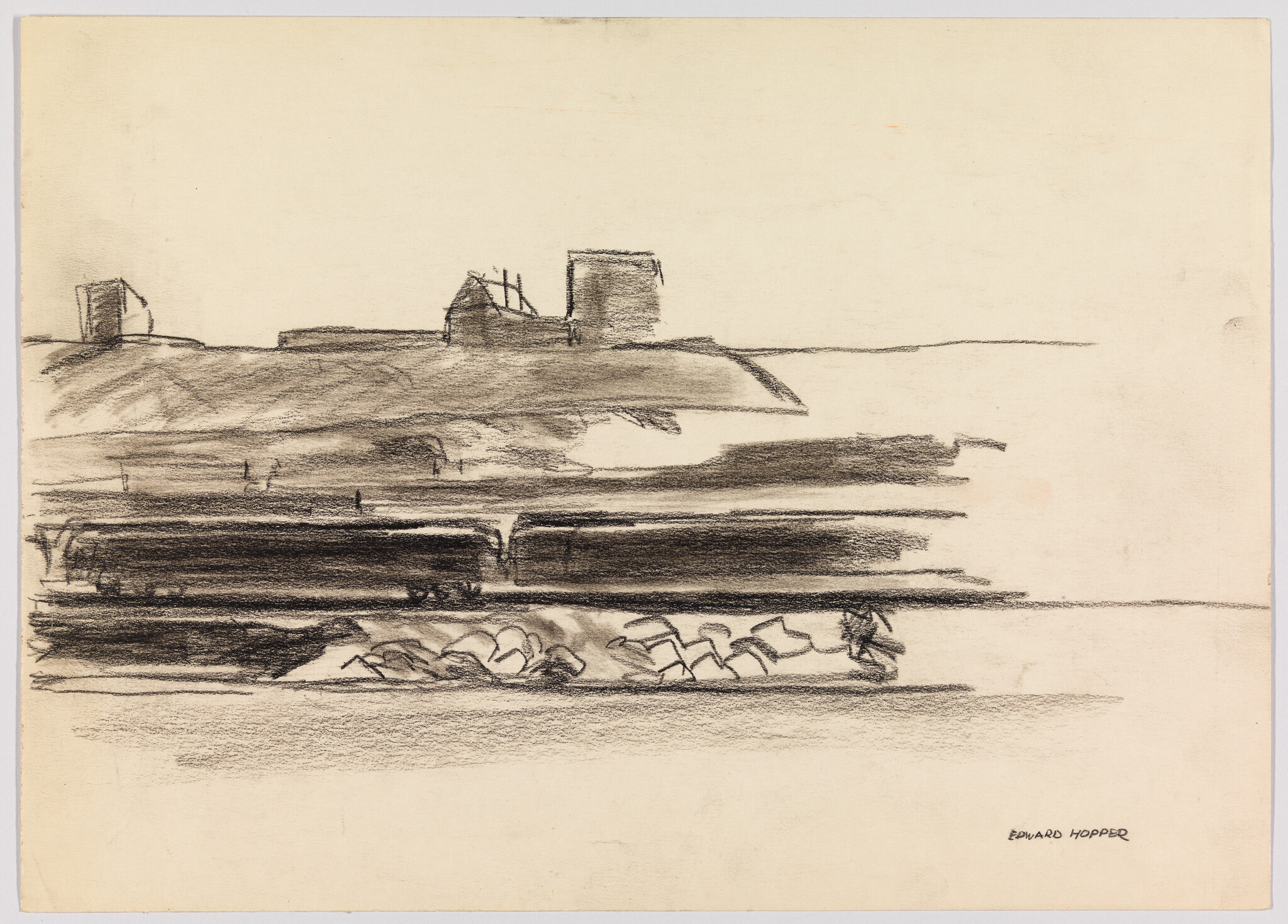 Freight train cars pass along tracks in front of a coastal hill with simple buildings.