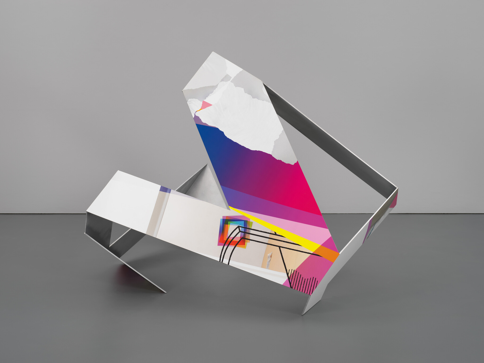 A modern art installation featuring a large, abstract geometric sculpture with mirrored surfaces and colorful graphic elements, displayed in a gallery with a gray floor and wall background.