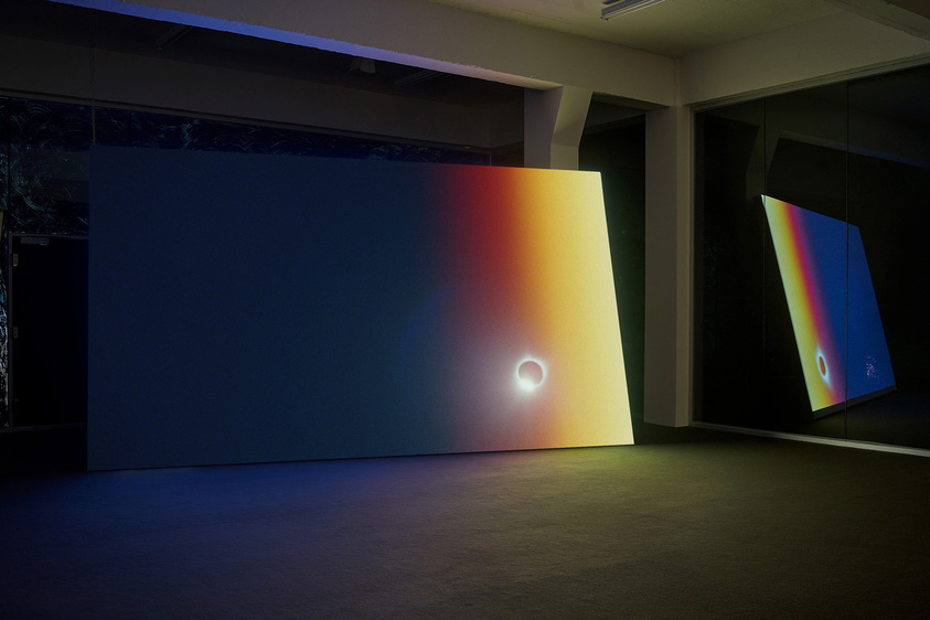 A dark room illuminated by two large panels displaying gradient colors and light patterns resembling a solar eclipse and its spectrum. The panel on the left shows a bright eclipse with a gradient from yellow to deep blue, while the right panel reflects a portion of the spectrum with a smaller eclipse image.