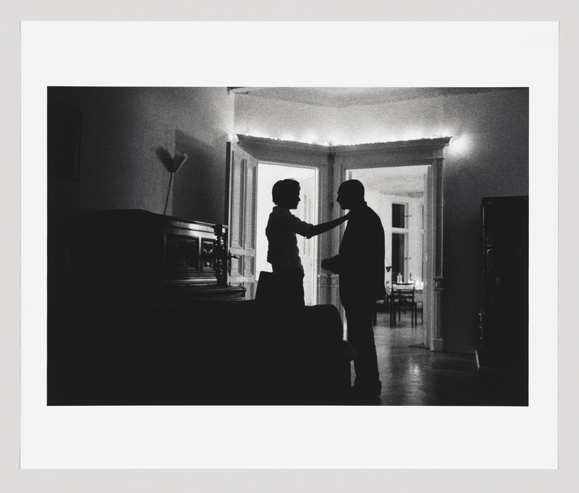 A black and white photo capturing a silhouette of a couple dancing in a dimly lit room with visible furniture outlines, including a piano, and a doorway leading to another room.