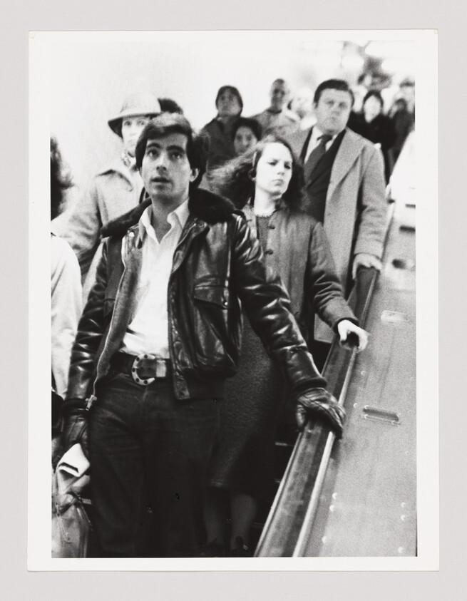 Man in leather jacket looks up while standing on a crowded escalator with other commuters.
