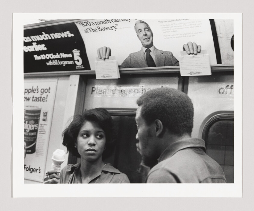 Young woman holding an ice cream cone looks at a man speaking to her on a subway.