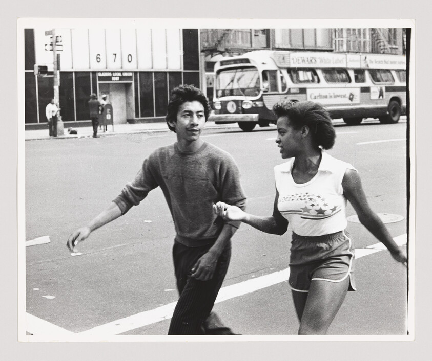 Young man and woman holding hands as they cross a city street, a bus passes behind them.