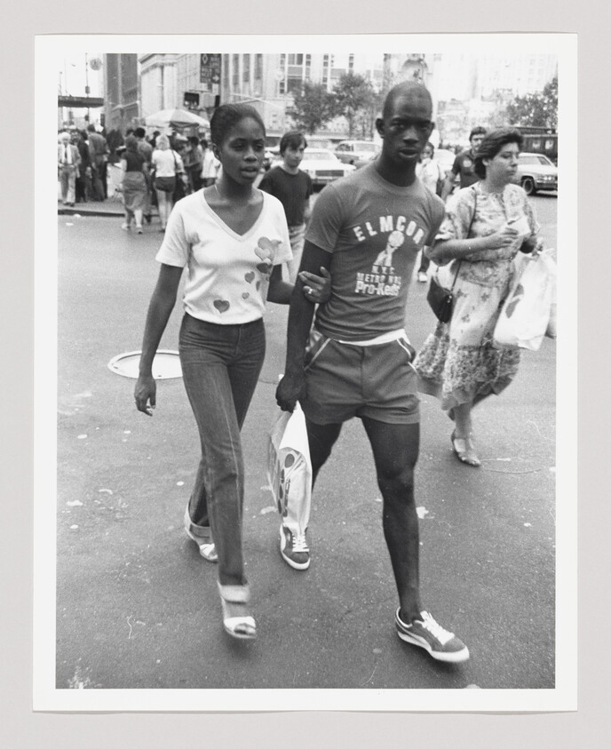 A young couple walks arm-in-arm through a busy city street carrying a shopping bag.
