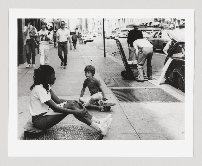 Two children sit on skateboards and talk on a city sidewalk while pedestrians pass by.