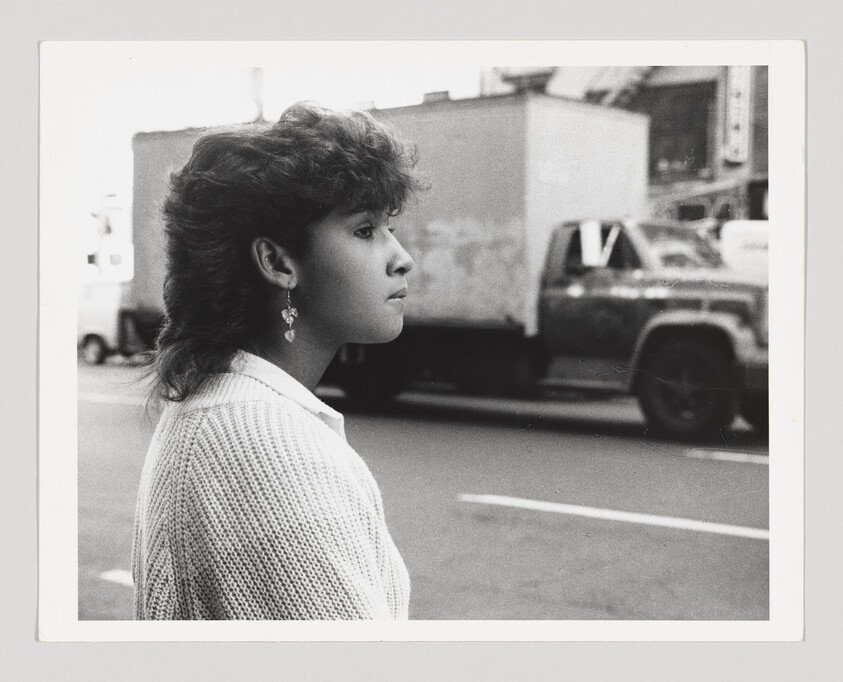 Young woman standing on a city street, looking to the right as a truck passes behind her.