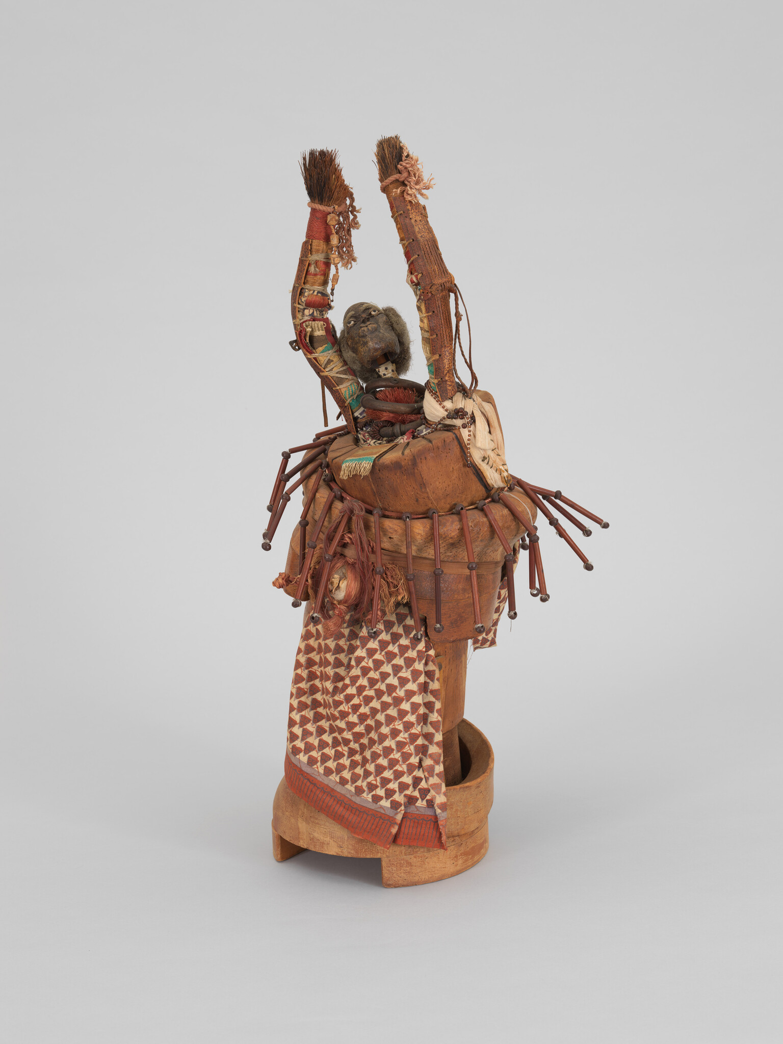 Carved wooden figure with raised arms wearing patterned cloth and bead and fiber decorations.