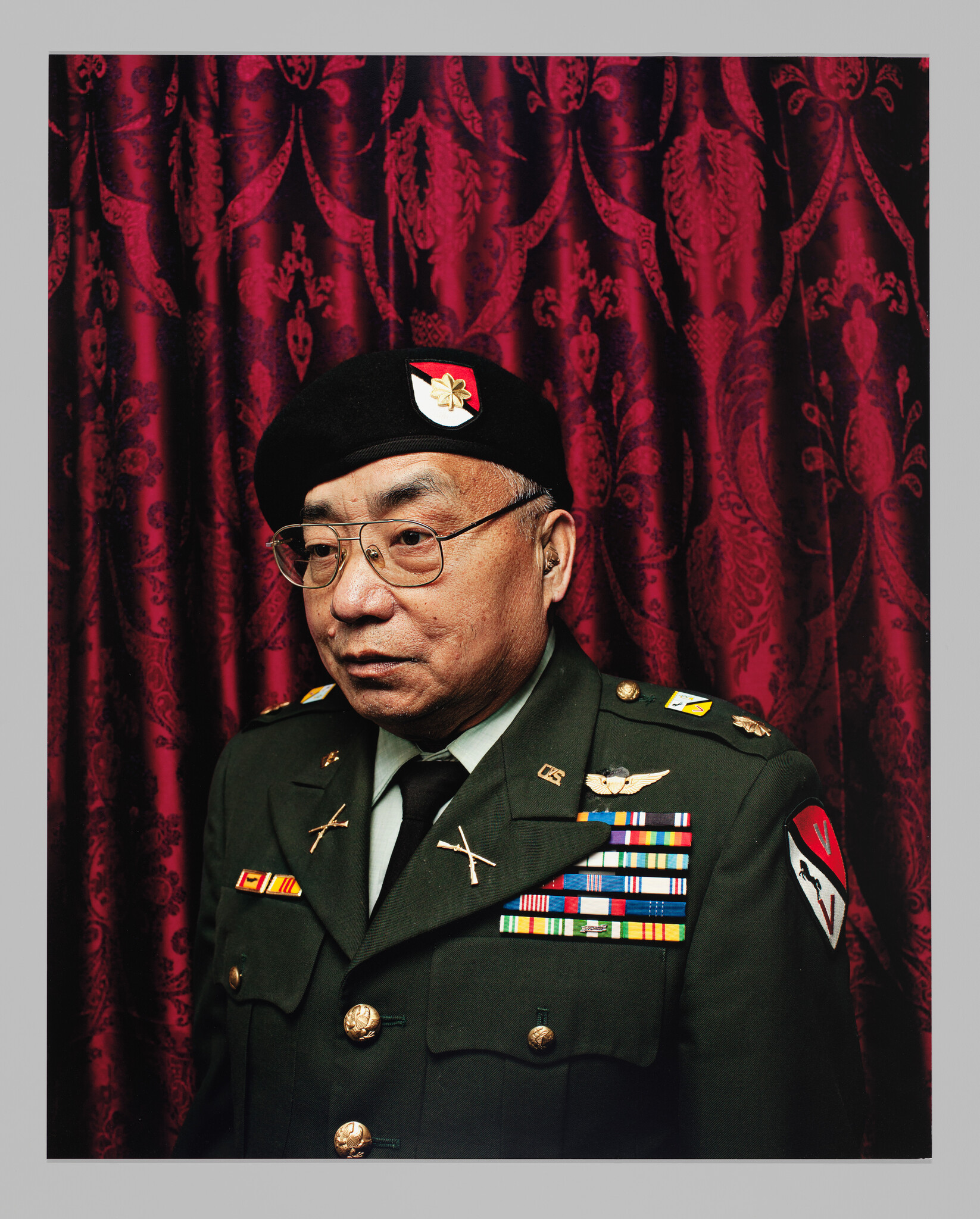 An older male veteran in a decorated military uniform and beret standing in front of a red curtain.