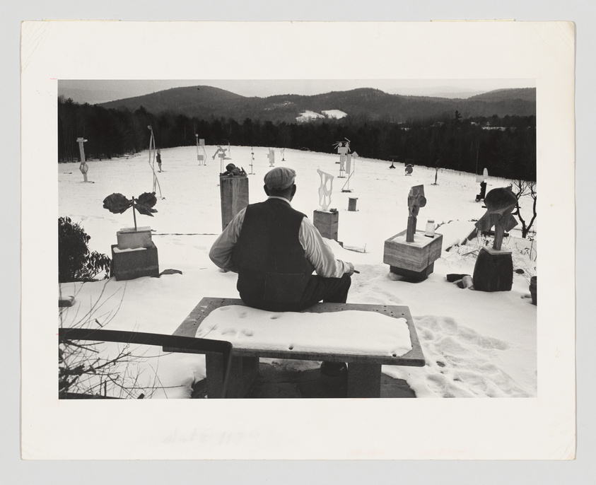 A person sits on a bench with their back to the camera, looking out over a snowy landscape dotted with sculptures and trees, with hills in the distance. The scene appears serene and contemplative.