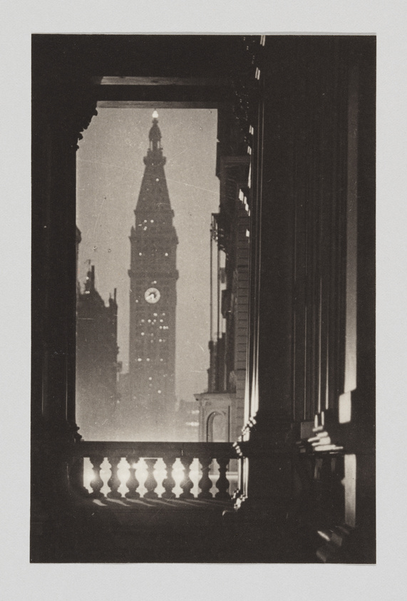 A vintage black and white photo depicting a view of a clock tower through a window with a decorative balustrade, framed by dark interior shadows. The image has a grainy texture and shows signs of age with scratches and specks on the surface.