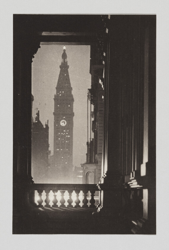 Tall clock tower framed by a dark building balustrade and archway at night.
