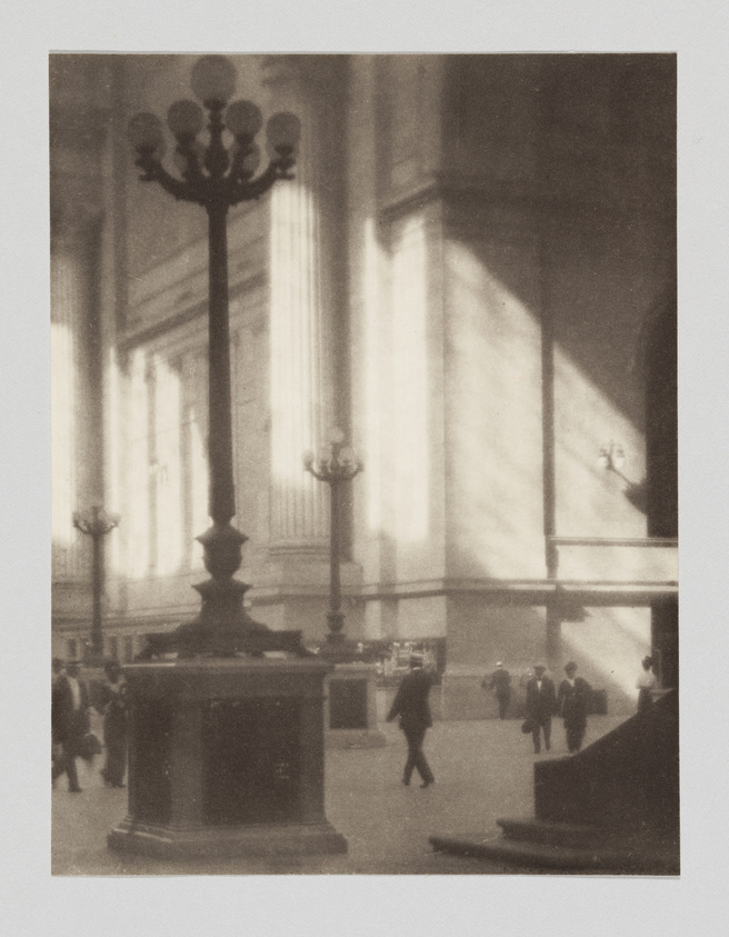 A sepia-toned photograph capturing the interior of a grand building with tall columns and large windows casting dramatic shadows. In the foreground, a prominent ornate candelabra stands, while in the background, several people appear to be walking or standing, engaged in various activities. The atmosphere is serene and steeped in history.