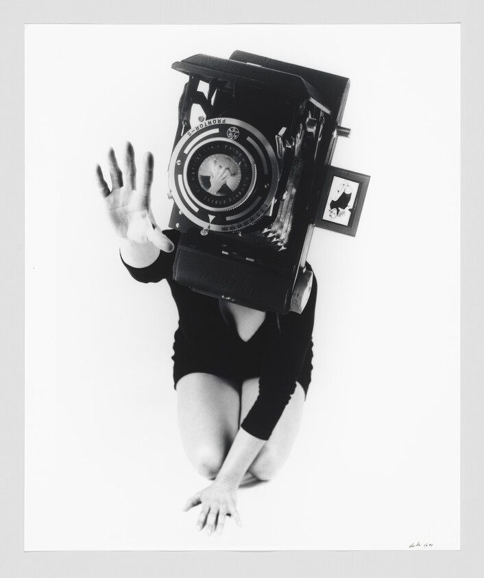Person kneeling with a large vintage camera covering their head, reaching a hand toward the viewer.