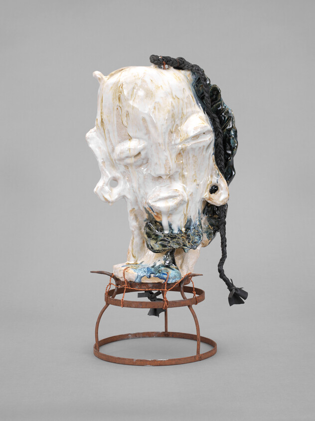 Sculpture of a distorted human head with closed eyes, braided hair, and a textured surface, mounted on a rusted metal stand.