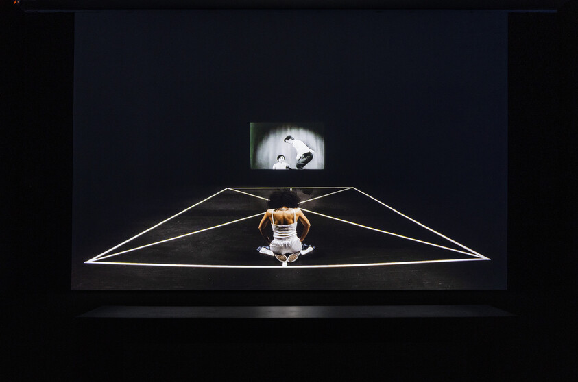 A person kneels inside glowing geometric lines on a dark stage while a black-and-white video plays.