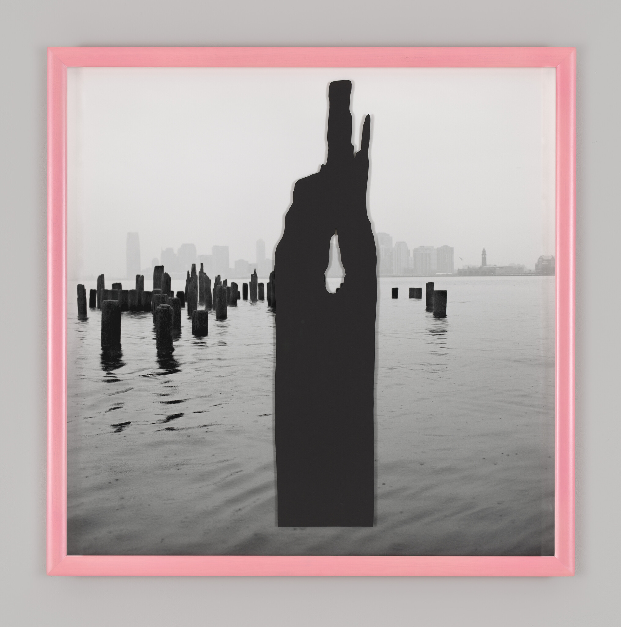 Large black sculptural silhouette stands in waterfront with old pilings and a faint city skyline.