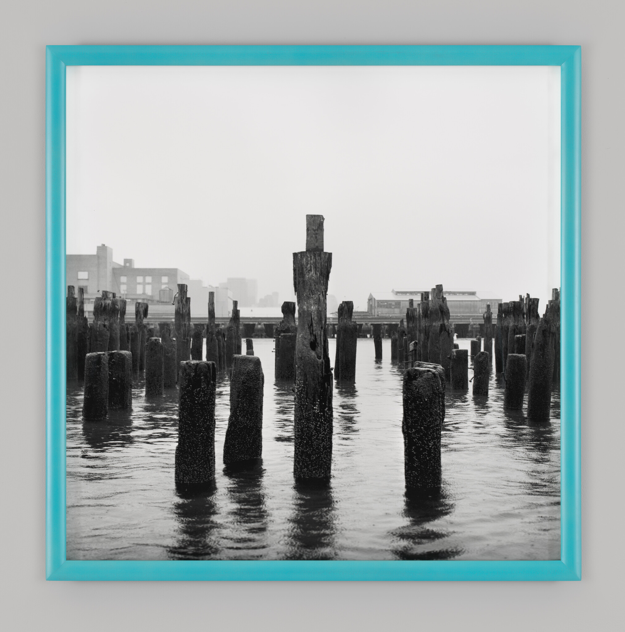 Weathered wooden pilings rise from calm water in front of distant waterfront buildings.
